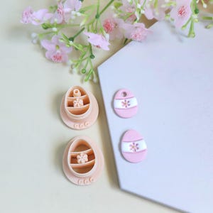 Peut inclure: Ensemble d'articles roses sur le thème de Pâques. On trouve deux emporte-pièces en forme d'œuf avec des détails floraux et deux ornements décoratifs en forme d'œuf avec des rayures blanches et des motifs floraux. Des fleurs roses sont en arrière-plan.