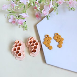 Könnte beinhalten: Gelbe ohrringförmige Ohrringe und rosa Ton-Ausstecher in Wabenform. Die Ohrringe haben eine Schlaufe zum Aufhängen. Die Ausstecher werden verwendet, um Ton in Wabenform zu formen. Im Hintergrund rosa Blüten.