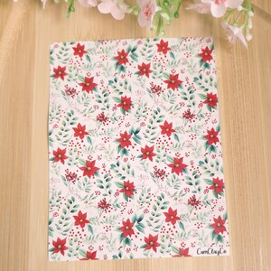 Peut inclure: Une feuille rectangulaire avec un motif répété de fleurs de poinsettia rouges, de feuilles vertes et de baies rouges sur un fond blanc. La feuille est posée sur une surface en bois brun clair. Le texte "CamClayCo" est visible.