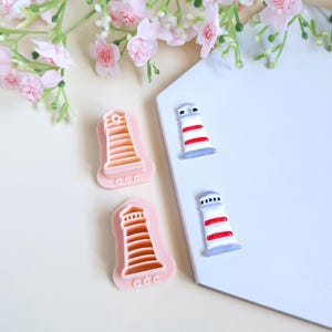 Peut inclure: Emporte-pièces roses en forme de phare et deux décorations en forme de phare. Les emporte-pièces ont un design détaillé, et les décorations présentent des rayures rouges et blanches. Le fond comprend des fleurs roses et une surface blanche.