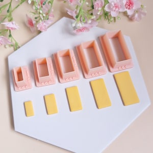May include: Set of six pink plastic rectangle-shaped moulds with measurements in millimetres. The moulds are arranged in order of size, with the smallest mould on the left and the largest mould on the right.  Below the moulds are five yellow rectangles of fondant.