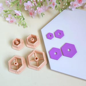 Puede incluir: Cuatro cortadores de arcilla hexagonales de color melocotón con punzones circulares centrales, etiquetados con los tamaños 1.5, 2.0, 2.5 y 3.0. Junto a ellos hay cuatro formas hexagonales de arcilla morada con agujeros centrales.