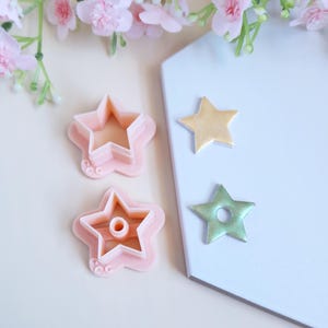 May include: Two pink plastic star-shaped cookie cutters with a hole in the center. A gold star and a green star are shown on a white surface.
