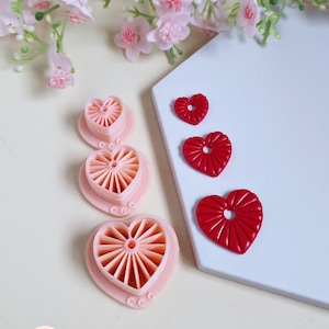 Op de afbeelding: Een verzameling hartvormige koekjessnijders en rode hartvormige versieringen. De koekjessnijders zijn lichtroze met een gedetailleerd interieurontwerp. De versieringen zijn felrood.