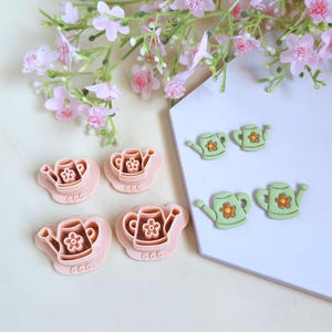 Peut inclure: Ensemble de fournitures artisanales en forme d'arrosoirs roses et verts. Les roses sont des emporte-pièces et les vertes sont des embellissements. Des fleurs roses sont en arrière-plan.