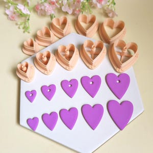 May include: A collection of peach-colored heart-shaped plastic clay cutters arranged in a semi-circle above a set of ten purple heart-shaped clay cutouts of varying sizes, some featuring small circular holes, displayed on a white hexagonal tile.