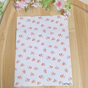May include: A white card with a pattern of orange and white clownfish and small bubbles. The card has the text "CamClayCo". Pink flowers and greenery are in the background. The card is on a wooden surface.