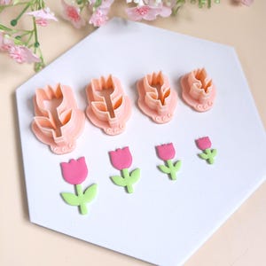 Könnte beinhalten: Satz mit vier rosafarbenen Tulpen-Ausstechern aus Kunststoff und drei rosafarbenen und grünen Fondant-Tulpen. Die Ausstecher sind in verschiedenen Größen und haben ein detailliertes Design.