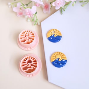 May include: Pink plastic cookie cutters featuring a sun and wave design, with two finished clay pieces of the same design. The clay pieces have a yellow sun and blue waves. The image includes pink flowers.