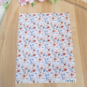 Puede incluir: Una hoja de papel estampada con un diseño repetido de fantasmas blancos, flores naranjas y hojas azules sobre un fondo crema. El texto "CamClayCo" es visible en la esquina inferior derecha. Elementos florales decorativos en el fondo.