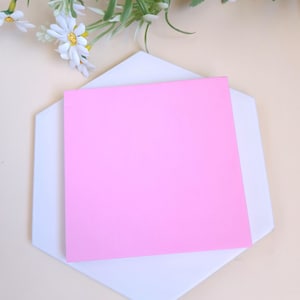 Peut inclure: Un carré de papier rose vif repose sur un dessous de plat hexagonal blanc. Des marguerites blanches artificielles avec des centres jaunes et des feuilles vertes sont disposées en haut. Le fond est de couleur beige clair.