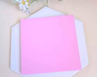 May include: A bright pink square of paper rests on a white, hexagonal coaster. Artificial white daisies with yellow centers and green leaves are arranged at the top of the image. The background is a light beige color.