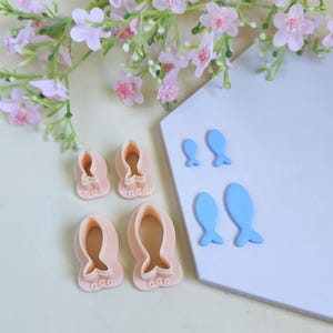 May include: A collection of pink and blue polymer clay cutters in the shape of fish. The cutters are in various sizes and are arranged on a white surface. Pink floral accents are in the background.