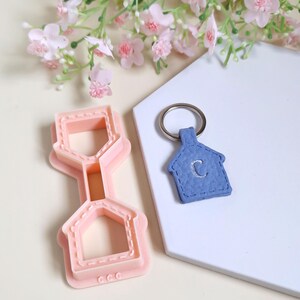 Peut inclure: Un emporte-pièce en forme de maison rose clair et un porte-clés en forme de maison bleu avec la lettre "C" brodée. Le porte-clés a un porte-clés argenté. Le fond comprend des fleurs roses et une surface blanche.