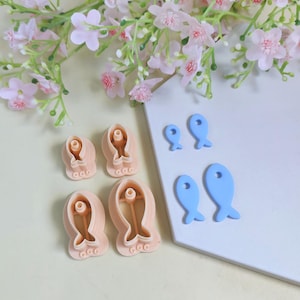 Puede incluir: Un conjunto de cortadores de galletas con forma de pez en varios tamaños y colores. Los cortadores son de color melocotón y azul. Flores rosas y vegetación en el fondo. La imagen está bien iluminada y es adecuada para listados de repostería.