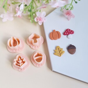Puede incluir: Un juego de seis cortadores de galletas y cortadores de fondant con temática otoñal. Los cortadores son de color rosa y presentan una calabaza, una bellota, un hongo y una hoja. Los cortadores de fondant tienen forma de calabaza, bellota, hongo y hoja. Los cortadores están sobre una superficie blanca.