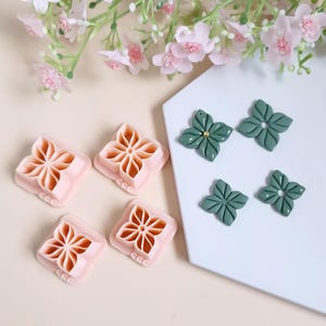 May include: Four pink plastic moulds shaped like a flower with a hole in the centre. Next to the moulds are two green polymer clay flowers with a gold dot in the centre.