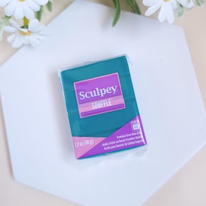 Pode incluir: Um bloco de argila polimérica Sculpey Soufflé turquesa, 48 g, em uma embalagem plástica transparente. O rótulo apresenta o nome da marca em branco e um detalhe roxo. O produto está sobre uma superfície geométrica branca, com flores brancas ao fundo.