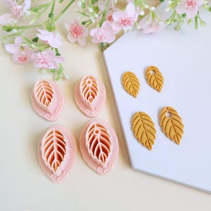 Könnte beinhalten: Eine Sammlung von blattförmigen Polymer Clay Ausstechern und fertigen Blattohrringen. Die Ausstecher sind rosa mit einem detaillierten Blattdesign, und die Ohrringe sind senfgelb. Der Hintergrund umfasst rosa Blüten und eine weiße Oberfläche.