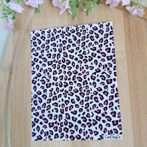 Peut inclure: Une feuille rectangulaire avec un motif léopard rose et noir sur fond blanc. La feuille est posée sur une surface en bois, avec des fleurs roses en arrière-plan. Le texte "CamClayCo" est visible en bas à droite.