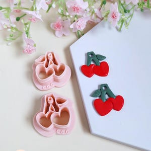 Peut inclure: Emporte-pièces roses en forme de cerises et deux boucles d'oreilles en pâte polymère en forme de cerises avec des cœurs rouges et des tiges vertes. Des fleurs roses sont en arrière-plan.