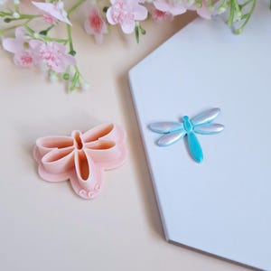 May include: A pink plastic dragonfly cookie cutter and a blue and silver dragonfly on a white background.