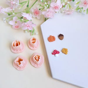 Peut inclure: Ensemble de découpoirs à biscuits roses en forme de citrouille et de feuille, avec des décorations miniatures sur le thème de l'automne. L'image est posée sur une surface blanche avec des accents floraux roses, créant un thème saisonnier.