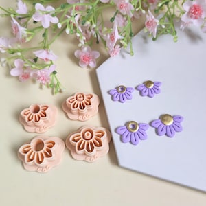 Peut inclure: Ensemble de découpoirs en pâte polymère en forme de fleurs et boucles d'oreilles assorties. Les découpoirs sont pêche, tandis que les boucles d'oreilles sont lavande avec des centres dorés. Des fleurs roses sont en arrière-plan.