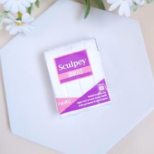 Peut inclure: Un paquet d'argile polymère blanche Sculpey Soufflé, 48 g, avec une étiquette rose et violette. Le paquet est sur une surface géométrique blanche, avec des fleurs de marguerite blanches en arrière-plan.