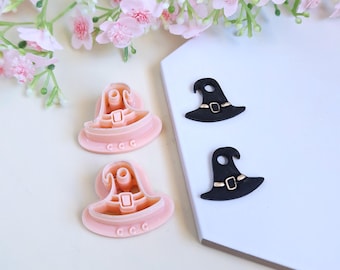 Op de afbeelding: Twee roze heksenhoedkoekjessnijders en twee zwarte heksenhoeddecoraties worden getoond op een wit oppervlak. De koekjessnijders hebben een gespontwerp. Roze bloemen staan op de achtergrond.