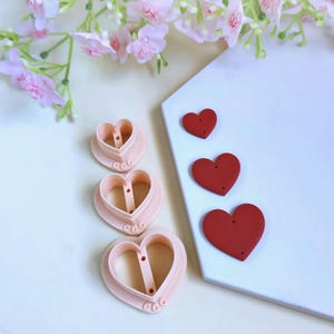 May include: Three heart-shaped cookie cutters and three red heart-shaped clay pieces are displayed on a white surface. The cookie cutters are light pink, and the clay hearts vary in size. Pink flowers are in the background.
