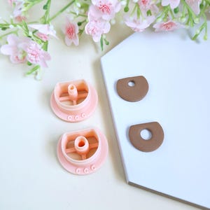May include: Two pink plastic cookie cutters with a central tube and three small circles, next to two brown, half-circle shaped pieces with a hole. Pink flowers are in the background.
