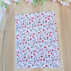 May include: A patterned card featuring a design of red hearts and dark green leaves on a white background. The card rests on a wooden surface, with pink flowers above. The text "CamClayCo" is visible.