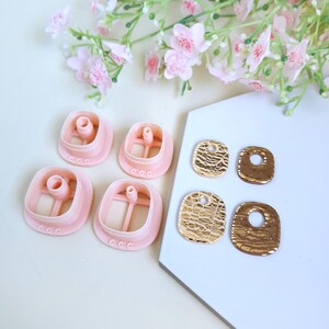 Puede incluir: Un conjunto de cortadores de galletas rosas cuadrados y pendientes dorados texturizados. Los cortadores de galletas tienen un tubo central y un peque&ntilde;o dise&ntilde;o decorativo. Los pendientes tienen forma rectangular con un agujero en la parte superior.