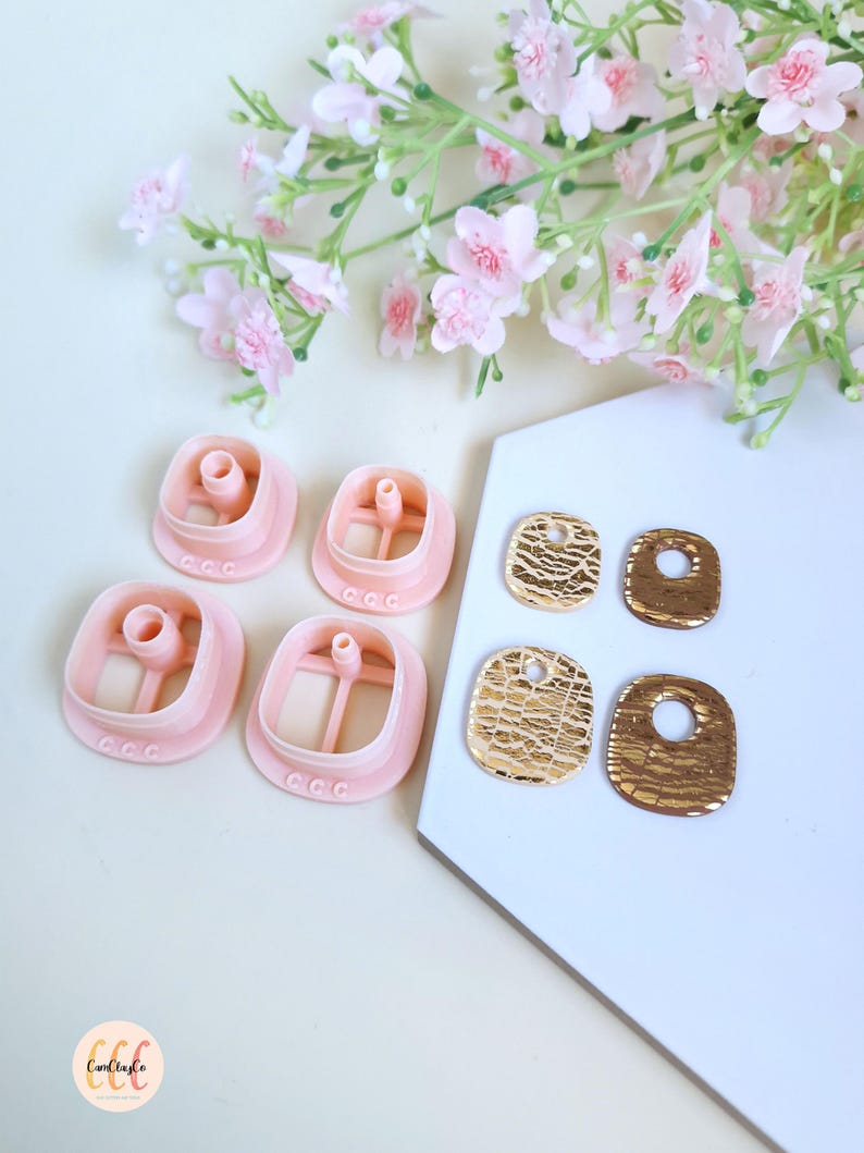 Puede incluir: Un conjunto de cortadores de galletas rosas cuadrados y pendientes dorados texturizados. Los cortadores de galletas tienen un tubo central y un peque&ntilde;o dise&ntilde;o decorativo. Los pendientes tienen forma rectangular con un agujero en la parte superior.