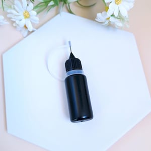 Peut inclure: Un petit flacon noir avec un embout compte-gouttes transparent, posé sur une surface hexagonale blanche. Le flacon est rempli d'un liquide foncé.