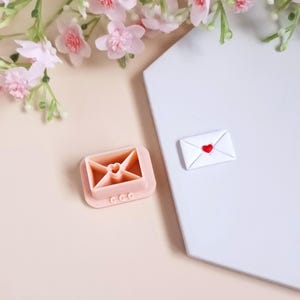 以下が含まれることがあります： ハートの切り抜きがあるピンクのプラスチック製の封筒型クッキーカッター。カッターは、赤いハートが描かれた小さな白い封筒のある白い表面の上に置かれています。