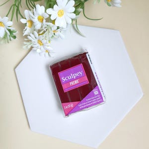 Peut inclure: Un paquet d'argile polymère Sculpey Premo, de couleur rouge foncé, repose sur une surface hexagonale blanche. L'emballage affiche le nom de la marque et les détails du produit. Des fleurs de marguerite blanches avec des centres jaunes sont disposées en arrière-plan. Le paquet contient 57 g d'argile.