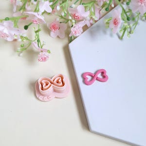 Peut inclure: Emporte-pièce rose en forme de nœud papillon, à côté d'une décoration rose en forme de nœud. Les objets sont sur une surface blanche, avec des fleurs roses en arrière-plan. Le découpoir a un design détaillé.