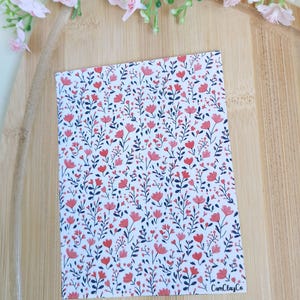 May include: A notebook with a floral pattern of red flowers and dark blue leaves on a white background. The rectangular notebook has rounded corners. The text "CamClayCo" is printed in the bottom right corner. Pink flowers are in the background.