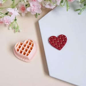 Peut inclure: Deux moules en silicone en forme de cœur, l'un avec un motif de gaufre et l'autre avec un design capitonné. Le moule à gaufres est rose clair et le moule capitonné est bordeaux avec des accents dorés. Les moules sont sur une surface blanche.