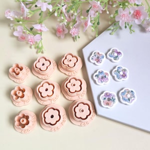 May include: A collection of pink flower-shaped clay cutters and floral-patterned embellishments. The cutters are arranged on a light surface, with a sprig of pink flowers in the background. The embellishments are on a white surface.