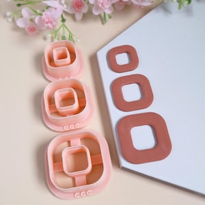 Peut inclure: Ensemble de quatre emporte-pièces carrés en plastique rose avec un carré plus petit découpé au centre. Les emporte-pièces sont disposés par ordre de taille, le plus grand en bas et le plus petit en haut. Les emporte-pièces sont sur une surface blanche.