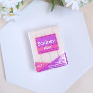 Puede incluir: Un paquete de 57 gramos de arcilla para modelar Sculpey Premo blanca. El paquete es morado y blanco con el texto "Sculpey Premo" en morado.