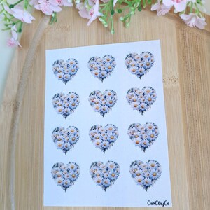 Könnte beinhalten: Ein Bogen weißer Aufkleber mit Gänseblümchen-Herz-Designs. Jedes Herz besteht aus weißen Blütenblättern und gelben Zentren, vor blauem Hintergrund. Die Aufkleber sind in einem Raster angeordnet. Der Markenname "CamClayCo" ist unten rechts aufgedruckt.