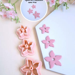 Puede incluir: Conjunto de cuatro cortadores de galletas en forma de flor rosa. Los cortadores están sobre una superficie blanca. El texto "Également disponible sans trou" es visible en la imagen.