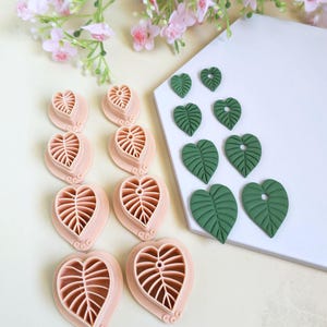 Puede incluir: Un juego de ocho cortadores en forma de hoja de corazón de color melocotón dispuestos en dos columnas, junto a ocho piezas de arcilla verde oscuro con forma de hoja con detalles de venas y pequeños agujeros en la parte superior.