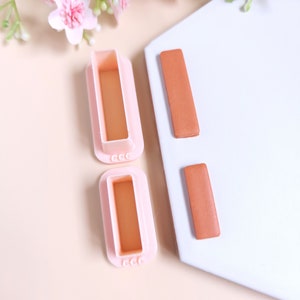May include: Two pink plastic molds for making rectangular shapes, and two brown clay rectangles. The molds have the letters "CCC" on the bottom.
