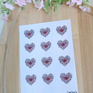 Könnte beinhalten: Ein Bogen weißer Aufkleber mit herzförmigen Blumendesigns. Jedes Herz besteht aus kleinen roten Rosen und winzigen weißen Blüten, umgeben von grünen Blättern. Die Aufkleber sind in Reihen und Spalten angeordnet. Der Markenname "CamClayCo" ist unten rechts aufgedruckt.