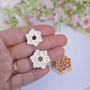 Könnte beinhalten: Zwei cremefarbene, blütenförmige Tonohrringe mit einem Spiraldesign werden in einer Hand gehalten. Ein passender Tonausstecher befindet sich in der Nähe. Die Ohrringe und der Ausstecher befinden sich auf einer weißen Oberfläche, mit rosa Blüten im Hintergrund.